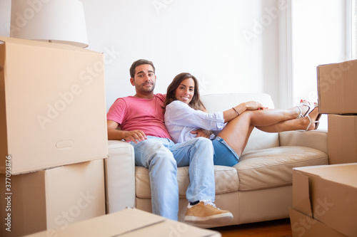 Happy Latin young couple sitting on couch among cardboard boxes in new house, looking at camera and smiling. Moving or relocation concept