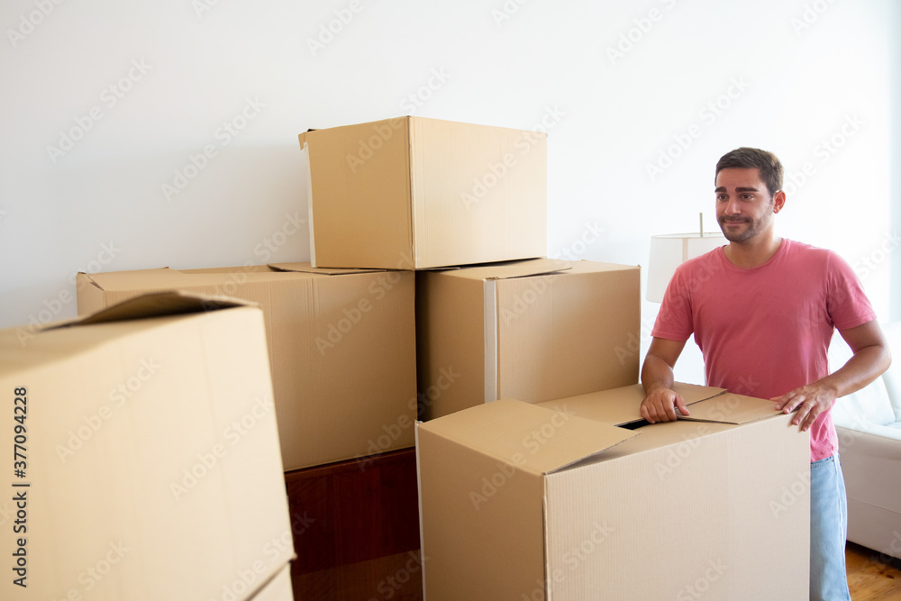 Fototapeta premium Happy young Hispanic man unpacking things in his new apartment, standing near stacks of carton boxes, looking at camera and smiling. Medium shot. Real estate concept