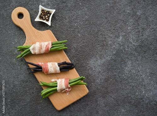 Prepared to bake appetizer, bundle of green beans wrapped in bacon on a wooden board on a dark concrete background. Hot appetizer recipes.