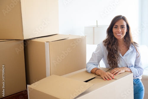 Happy beautiful young woman unpacking things in her new apartment, standing near stacks of carton boxes, looking at camera and smiling. Medium shot. Real estate concept