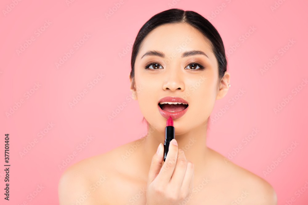 naked asian woman applying lipstick while looking at camera isolated on pink naked asian woman applying lipstick while looking at camera isolated on pink