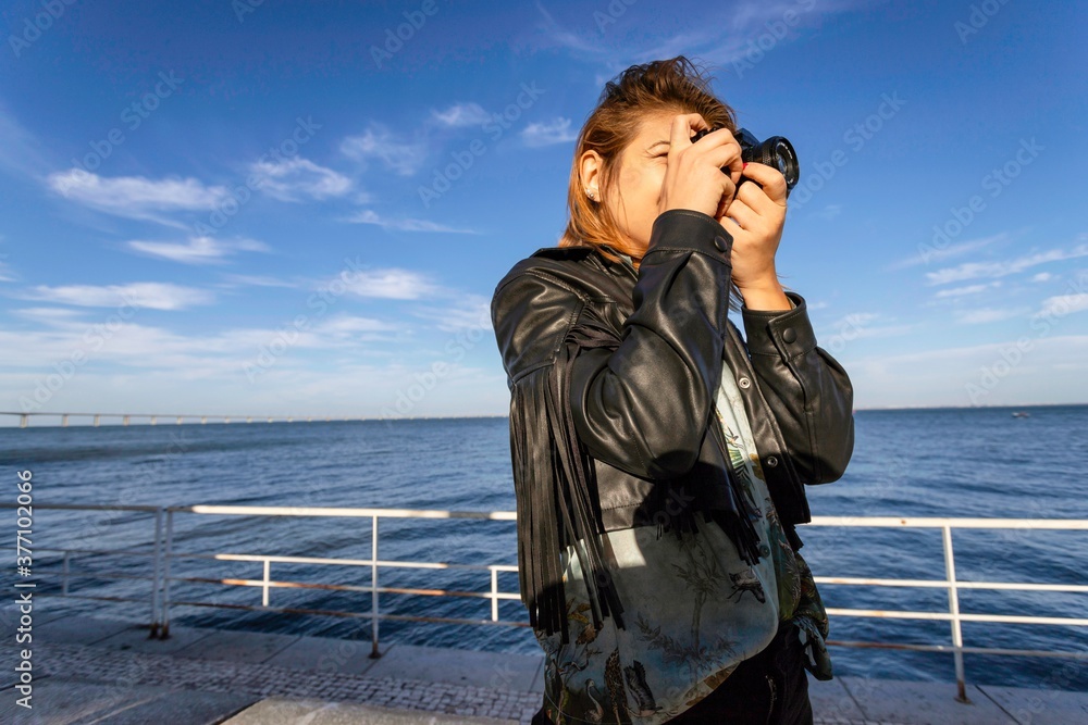 Young woman with blond hair black jacket, uses mobile phone for texting with friends, reads post online, connected to wireless internet, concentrated in display and take photos