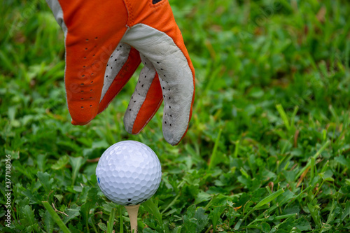 Close-up hand with orange glove and white golf ball on tee, on green grass, horizontally, with copy space