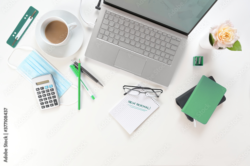 White office desk with laptop, green working utilities, surgical face mask against coronavirus and covid-19 and notepad with text New Normal, business concept, copy space, high angle view from above White office desk with laptop, green working utilities, surgical face mask against coronavirus and covid-19 and notepad with text New Normal, business concept, copy space, high angle view from above