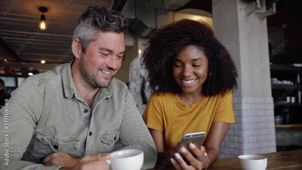 Good-looking couple laughing at social media post on smartphone drinking hot coffee while sitting n stylish cafe. Good-looking couple laughing at social media post on smartphone drinking hot coffee while sitting n stylish cafe.