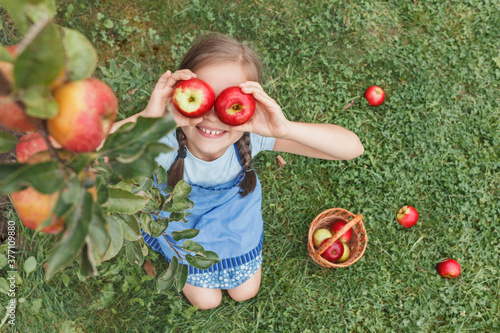 Apples for Children. Apples Eyes Kid. Apple Harvest. Little girl in blue apron put two apples to her eyes on grass background © Maryana