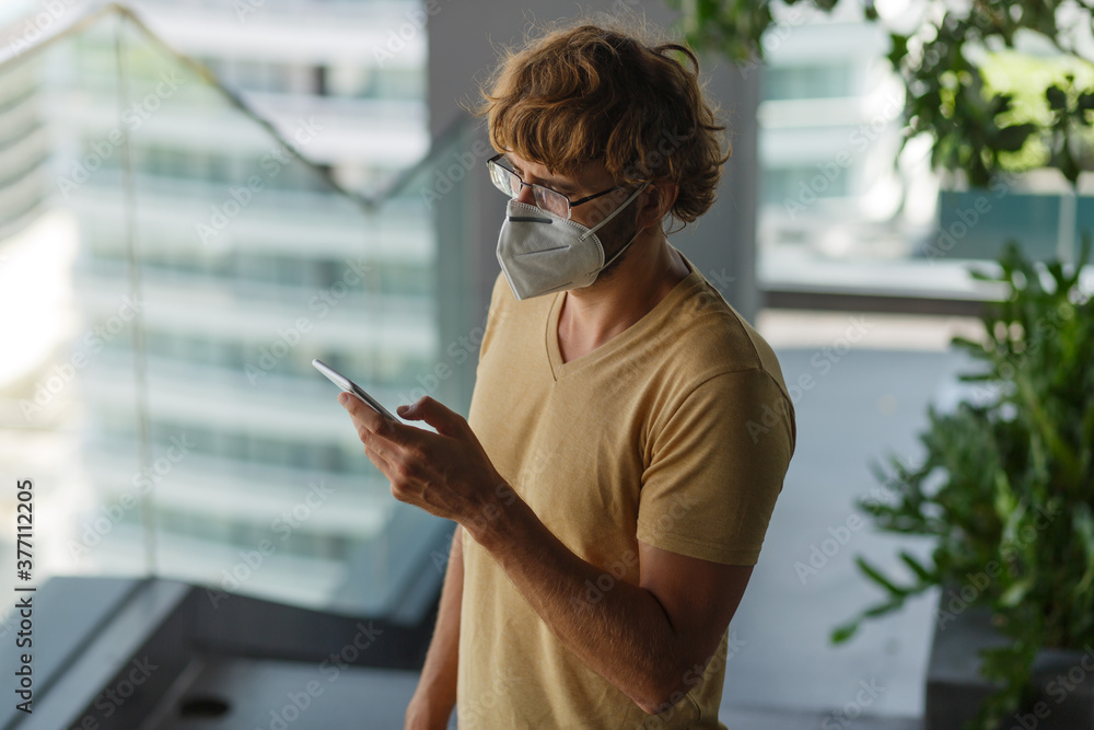 White bearded adult man using smartphone while wearing surgical mask on an industrial wall. Health, epidemics, social media.