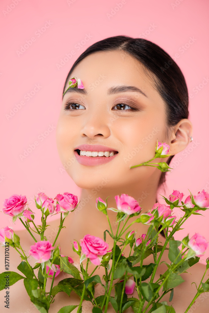 young asian woman with flowers on face looking away near tiny roses isolated on pink