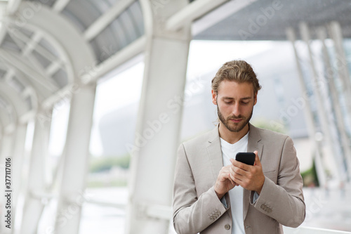 Portrait of handsome curly businessman in casual wear holding smartphone and smiling.Successful manager using mobile phone apps,texting message,browsing internet,looking at phone near business center.