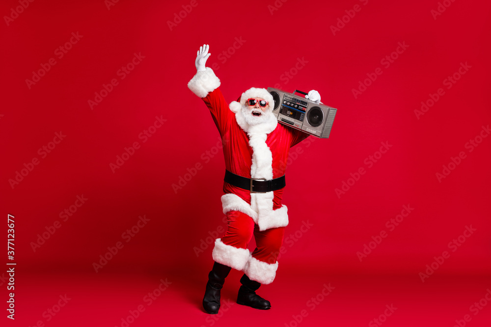 Full length photo of retired old man white beard careless raise hand hold listen retro radio tape wear x-mas santa costume gloves coat belt sunglass cap boot isolated red color background Full length photo of retired old man white beard careless raise hand hold listen retro radio tape wear x-mas santa costume gloves coat belt sunglass cap boot isolated red color background