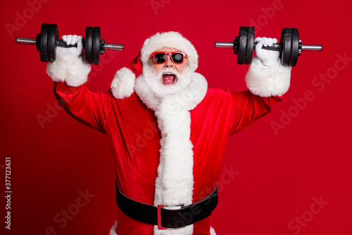 Photo of pensioner old man white beard lift heavy dumbbells smiling open mouth screaming get mad excited training wear santa costume sunglass headwear isolated red color background