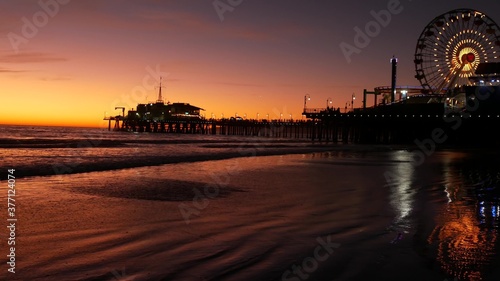 Twilight waves against classic illuminated ferris wheel, amusement park on pier in Santa Monica pacific ocean beach resort. Summertime iconic symbol of California glowing in dusk, Los Angeles, CA USA.