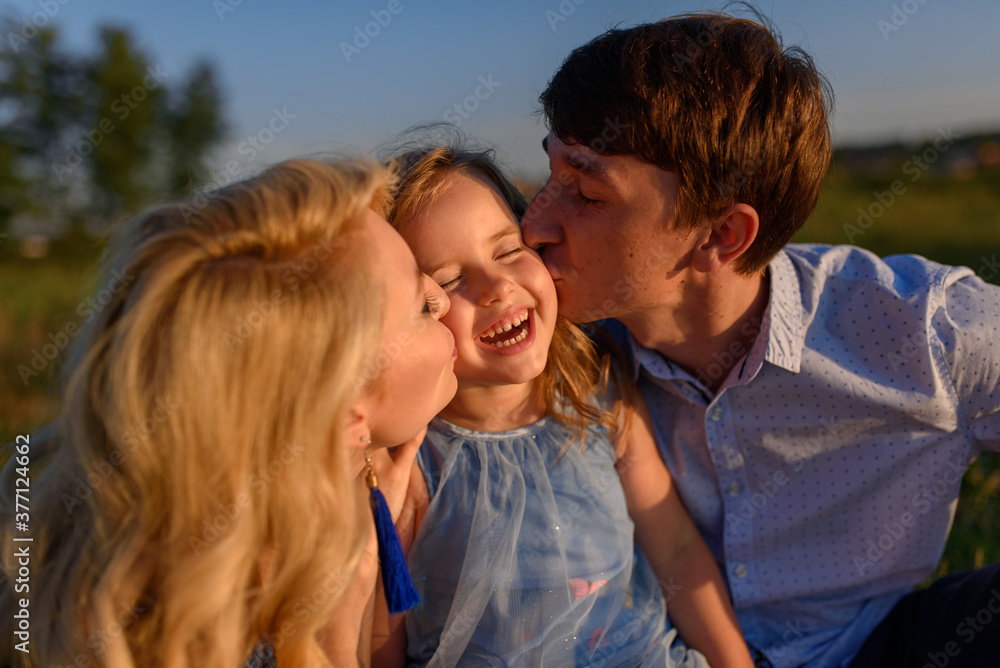 Dad mom and daughter are sitting together on the grass. Parents kiss their daughter on the cheeks. Dad mom and daughter are sitting together on the grass. Parents kiss their daughter on the cheeks.