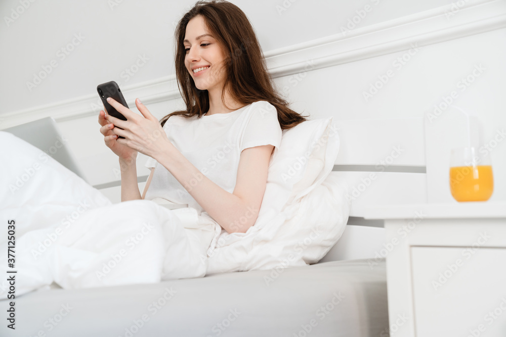 Image of cheerful girl using cellphone and laptop while sitting in bed Image of cheerful girl using cellphone and laptop while sitting in bed