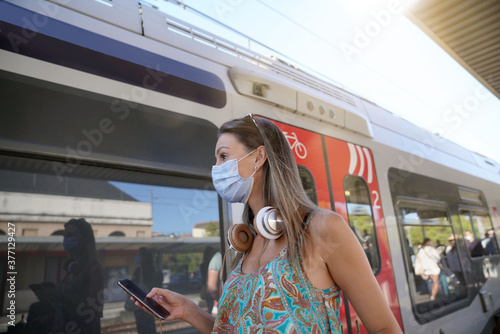 Young woman waiting for train on railway platform, wearing face mask