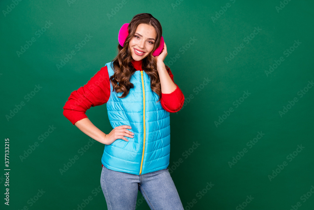 Photo of pretty cute young lady hands hip beaming shiny smiling warm cafe winter weather wear pink ear warmers jeans blue zipped vest red sweater isolated green color background