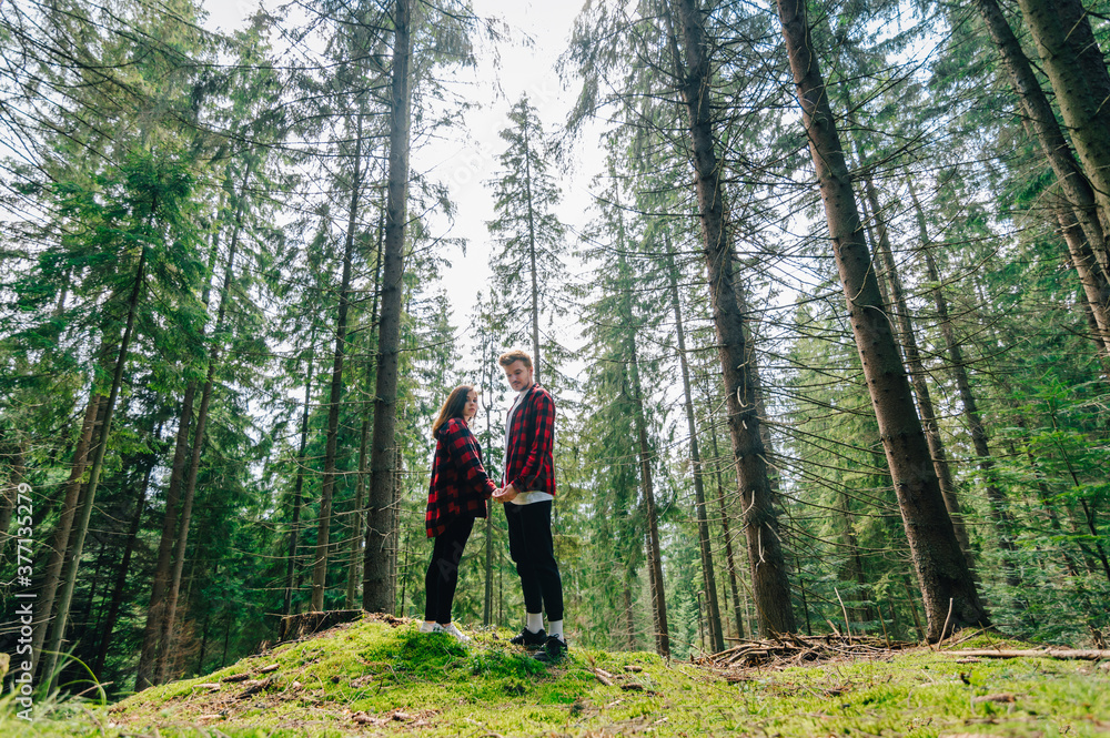 Young couple man and woman in red shirts stand in the woods on the moss, holding hands and posing for camera. Couple of hipsters on a hike in the woods holding hands on a background of beautiful trees