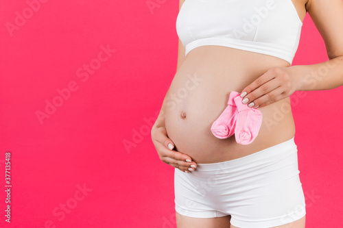 Close up portrait of pregnant woman in white underwear holding pink socks for a baby girl at pink background. Childbirth expecting. Copy space © sosiukin