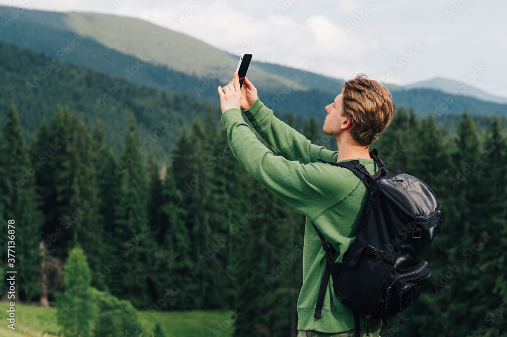 Young man in casual clothes catches a cellular network on a smartphone with his hands raised. Guy tourist takes a selfie on a hike against the backdrop of mountain forest and mountains.