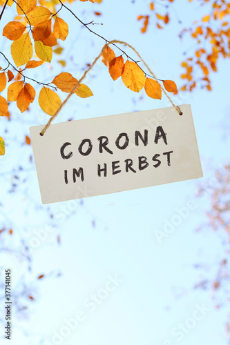 weißes Schild aus Holz mit Schriftzug: Corona im Herbst, hängt im Herbst, vor blauem Himmel, an einem Ast mit schönem gelben und orangen Herbslaub. © Barbara-Maria Damrau