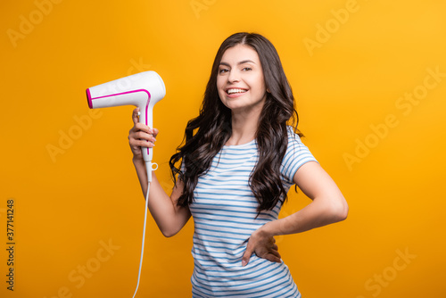 brunette woman with curls holding hairdryer isolated on yellow