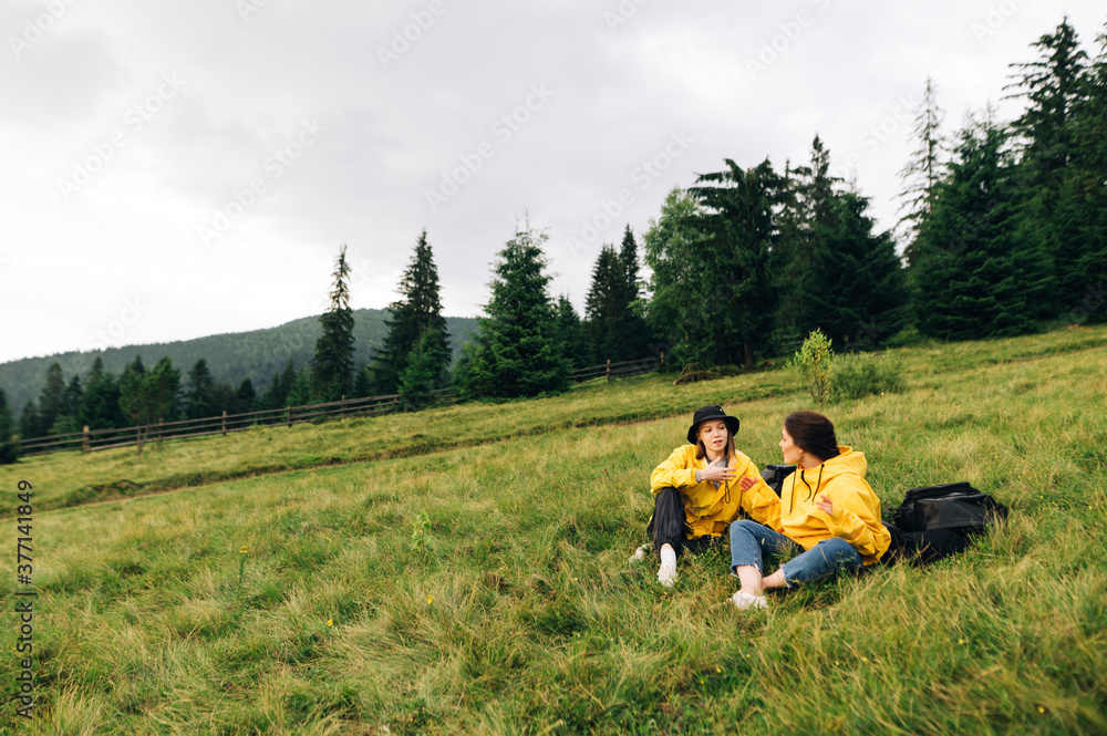Two girls tourists in yellow raincoats sitting on a meadow in the mountains against the backdrop of a beautiful landscape, relaxing and talking. Hikers rest on a meadow.