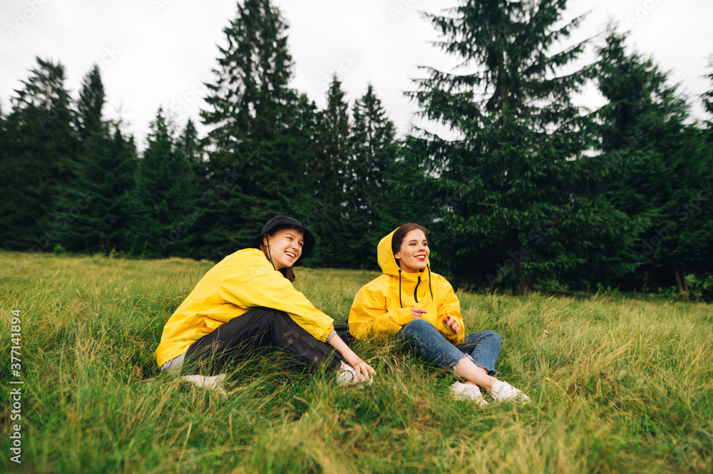 Positive girl tourist in yellow raincoats sitting on a bitter meadow and looks away with a smile on his face. Women on a hike rest on the grass, communicate with a happy face.