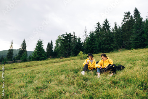 Two women tourists in yellow raincoats on a hike sat down to rest in a meadow. Two hikers in the mountains resting on a meadow on a background of forest. Mountain tourism