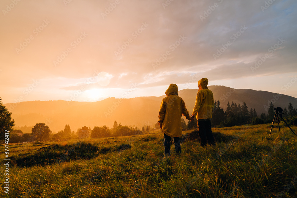 People in yellow raincoats stand on a meadow in the mountains and watch the beautiful sunset in the rain and shoot with a camera on a tripod. Background. Hikers in the rain at sunset on a hike