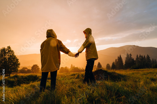 Couple of young hikers in yellow raincoats standing on a meadow in the mountains holding hands on a background of sunset. 2 people got into the evening rain at sunset in the mountains © Serhii