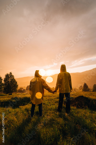 View from the back. Photo with highlights. Couple of young hikers standing in the rain holding hands in the mountains at sunset and looking at the unreal landscape.