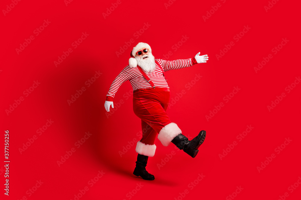Full length photo of grandfather grey beard go look camera wear santa claus x-mas costume suspenders sunglass striped shirt cap boots isolated red color background