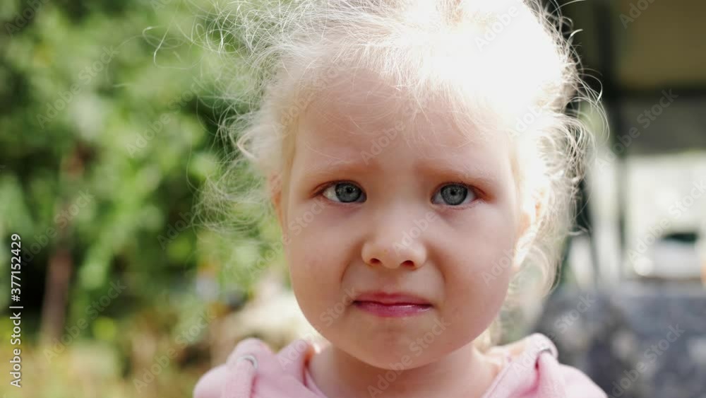 Little baby girl with blue eyes and curly hair looking at camera. Young serious child looking at the camera. Close Up. Outdoor. Portrait of sad little kid. Little baby girl with blue eyes and curly hair looking at camera. Young serious child looking at the camera. Close Up. Outdoor. Portrait of sad little kid.