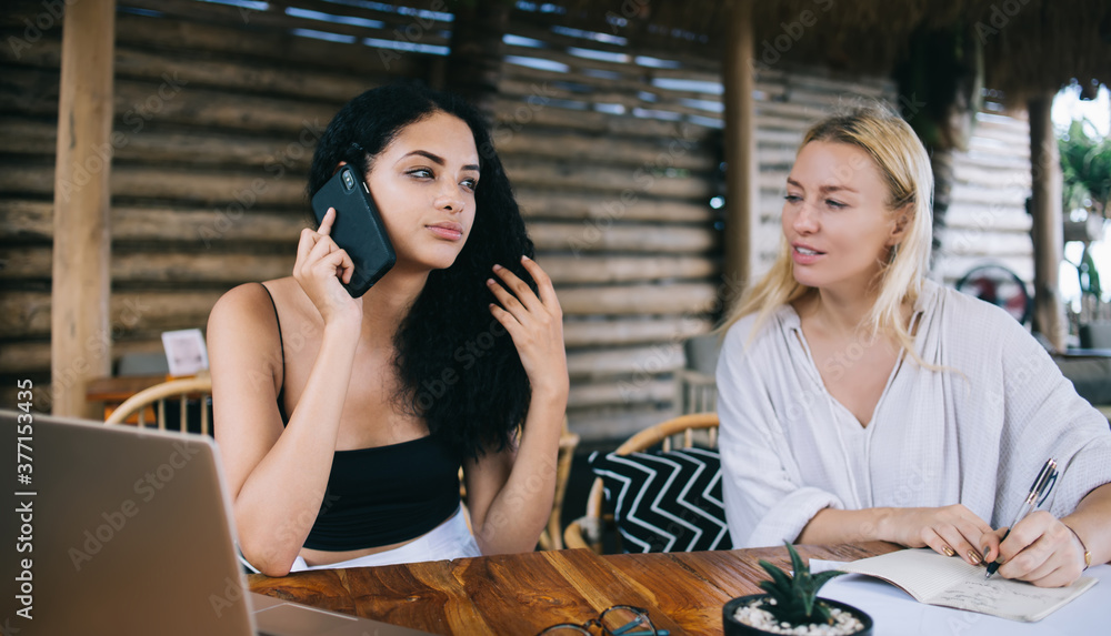 Young brunette female making call to service operator making booking for delivery while spending time with friend, caucasian women working remotely together using modern technology for research