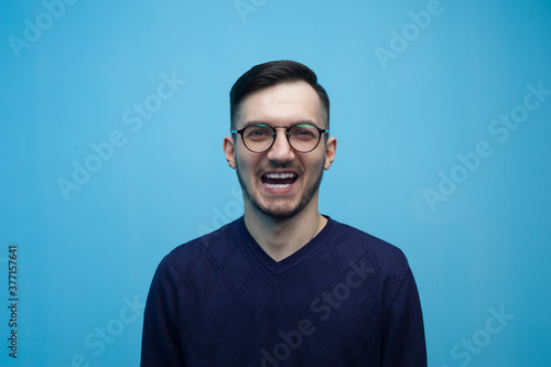 Portrait of an emotional young man in glasses shocked by low prices and discounts on a blue background. © pavel_shishkin