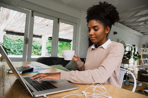 Female student making notes typing on laptop holding hot coffee sitting in fancy kitchen. photo