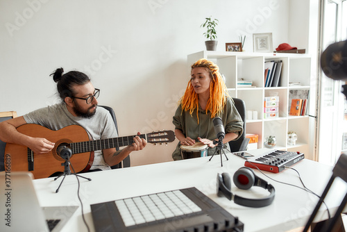 Man playing guitar and woman singing, playing rhythm with djembe drum while recording video blog or vlog. Couple of musicians making music at home