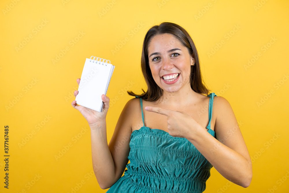 Young beautiful woman over isolated yellow background smiling, surprised and pointing blank notebook in her hand Young beautiful woman over isolated yellow background smiling, surprised and pointing blank notebook in her hand