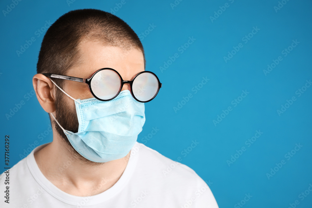 Man with foggy glasses caused by wearing disposable mask on blue background, space for text. Protective measure during coronavirus pandemic