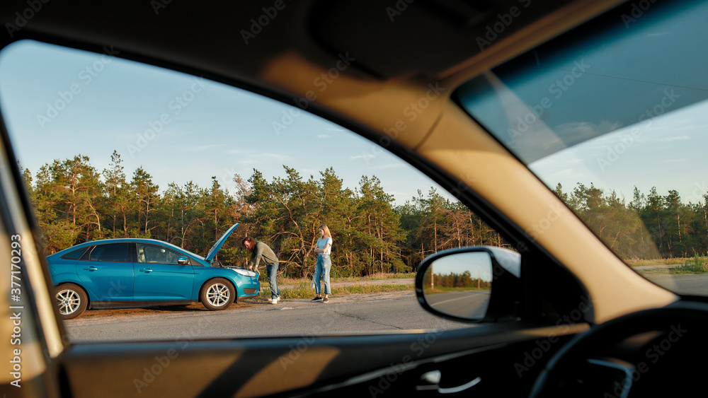 Obraz premium Couple having troubles with their auto, Man looking under the hood of broken car and trying to repair it while his girlfriend waiting for him, View from another car window