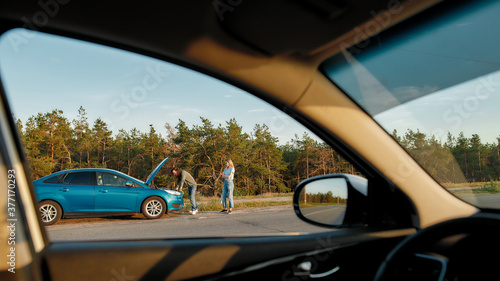 Couple having troubles with their auto, Man looking under the hood of broken car and trying to repair it while his girlfriend waiting for him, View from another car window © Svitlana