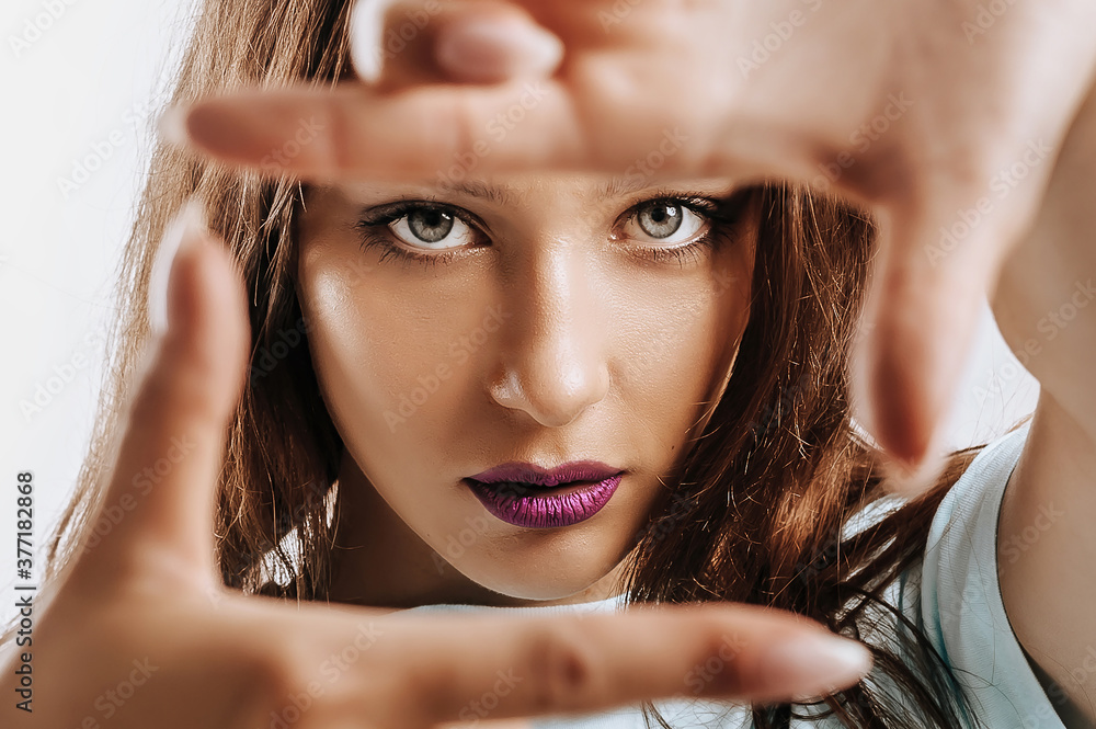 Beautiful young girl makes a frame with her hands on an isolated gray background. The woman with fashion make-up with purple lips seriously looking at the camera. Place for advertising.