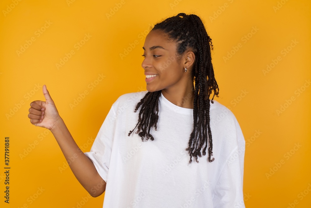 Young dark skinned woman with braids hair wearing white t-shirt over yellow background Looking proud, smiling doing thumbs up gesture to the side. Good job! Young dark skinned woman with braids hair wearing white t-shirt over yellow background Looking proud, smiling doing thumbs up gesture to the side. Good job!