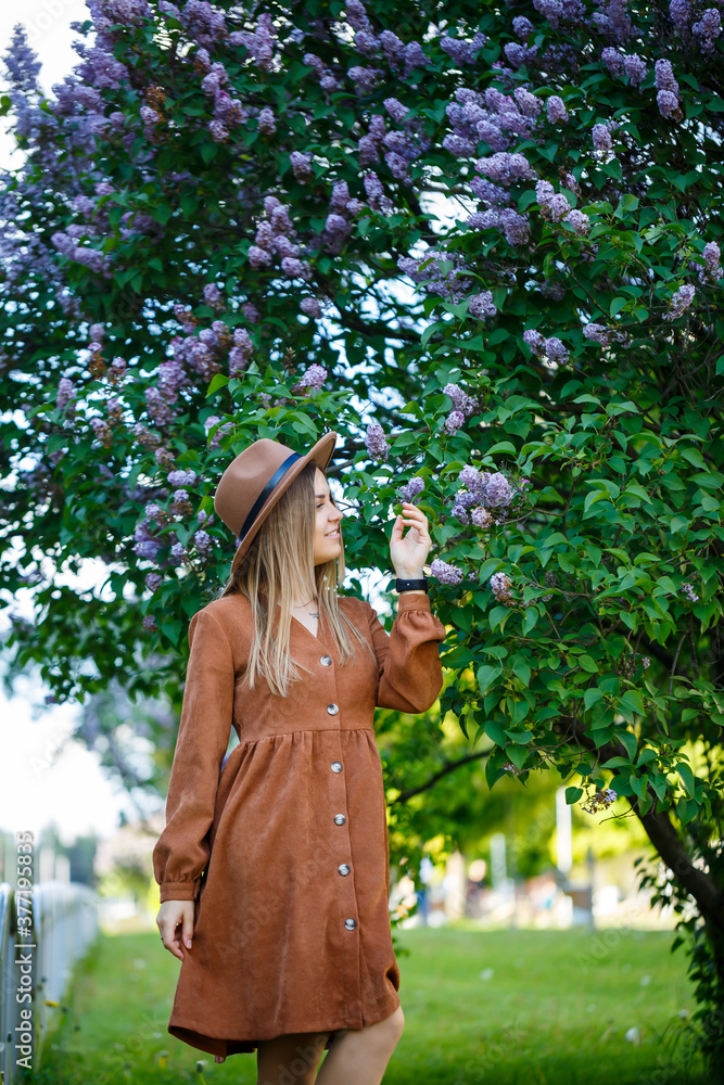 Portrait of a stylish girl in a brown hat and dress on a background of lilacs tree on a sunny warm day. A young woman of European appearance with a smile on her face