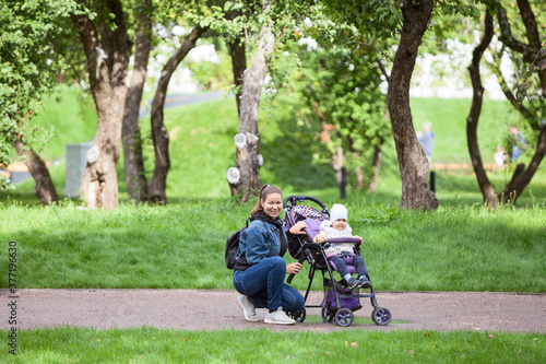 Mother and her toddler daughter walking in garden with infant stroller, woman sitting close to girl, horizontal image