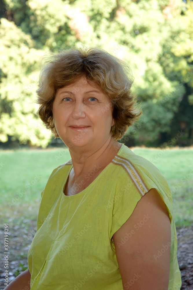 Vertical portrait of mature confident lady in green blouse looking at camera and smiling in the park, blurred background Vertical portrait of mature confident lady in green blouse looking at camera and smiling in the park, blurred background