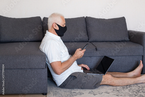Online job concept. Serious elderly man in medical mask working at home on his laptop computer and using smartphone