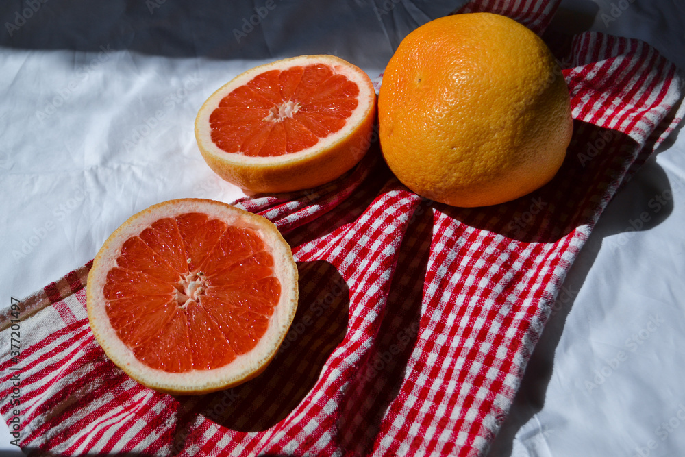two halves cut red orange grapefruit and whole on white cloth and red fabric with folds in light of sun with shadows two halves cut red orange grapefruit and whole on white cloth and red fabric with folds in light of sun with shadows