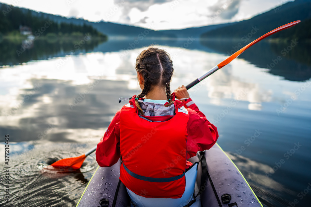Caucasian woman in a kayak enjoys the clear view in front of her, with pine forest reflected in the beautiful and calm lake on a beautiful day of wild adventure. Hobby and leisure activity in nature. Caucasian woman in a kayak enjoys the clear view in front of her, with pine forest reflected in the beautiful and calm lake on a beautiful day of wild adventure. Hobby and leisure activity in nature.