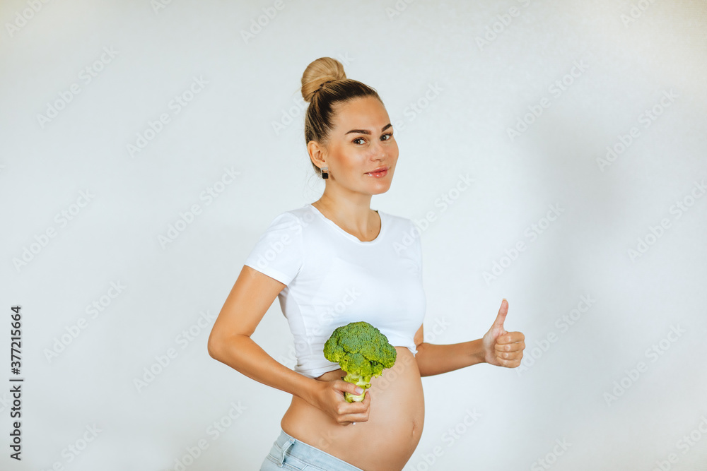 Pretty young pregnant woman in white shirt and jeans posing with fresh broccoli on white background. Pretty young pregnant woman in white shirt and jeans posing with fresh broccoli on white background.